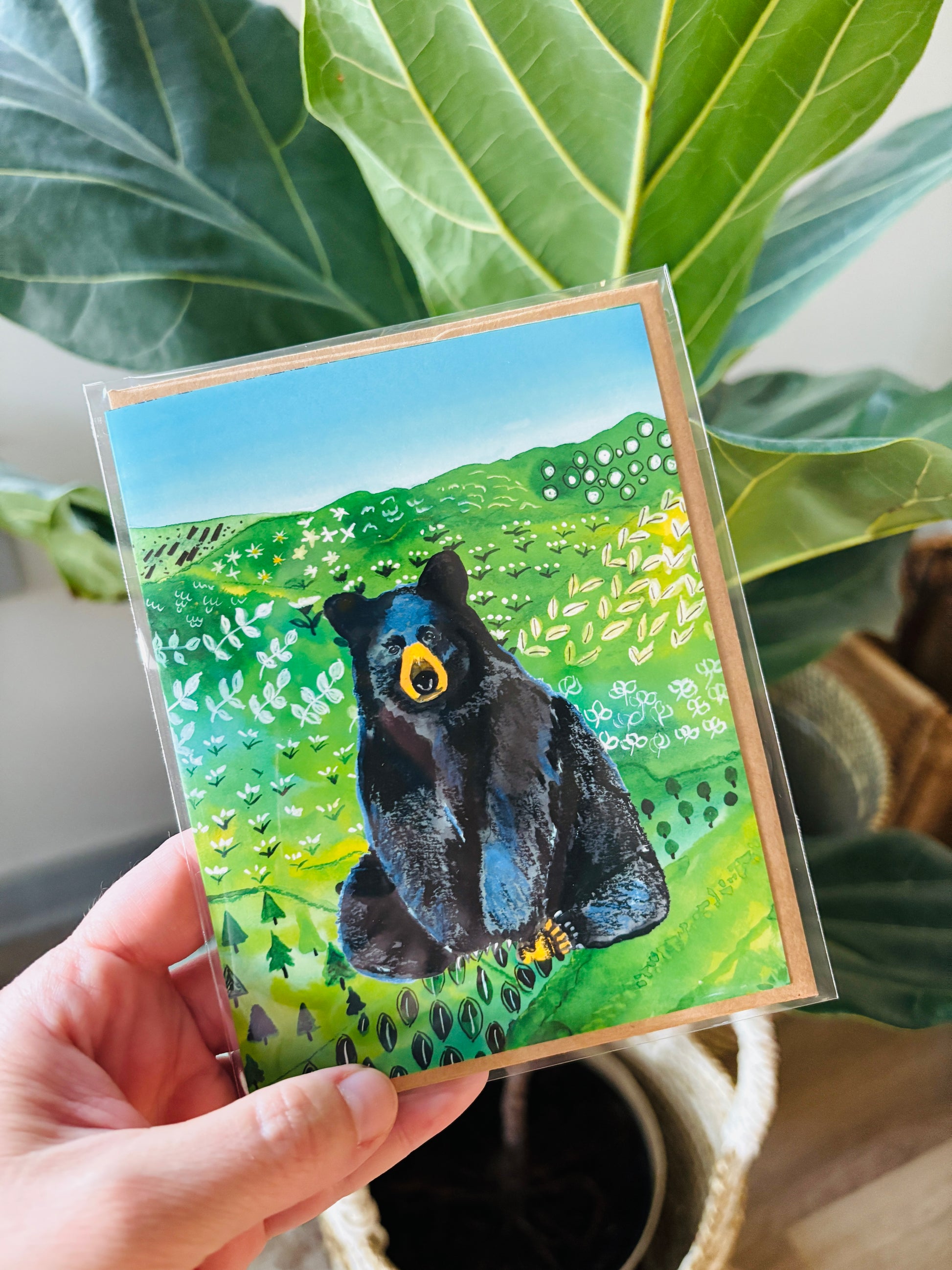 Hand holding a card with a bear illustration in a natural setting
