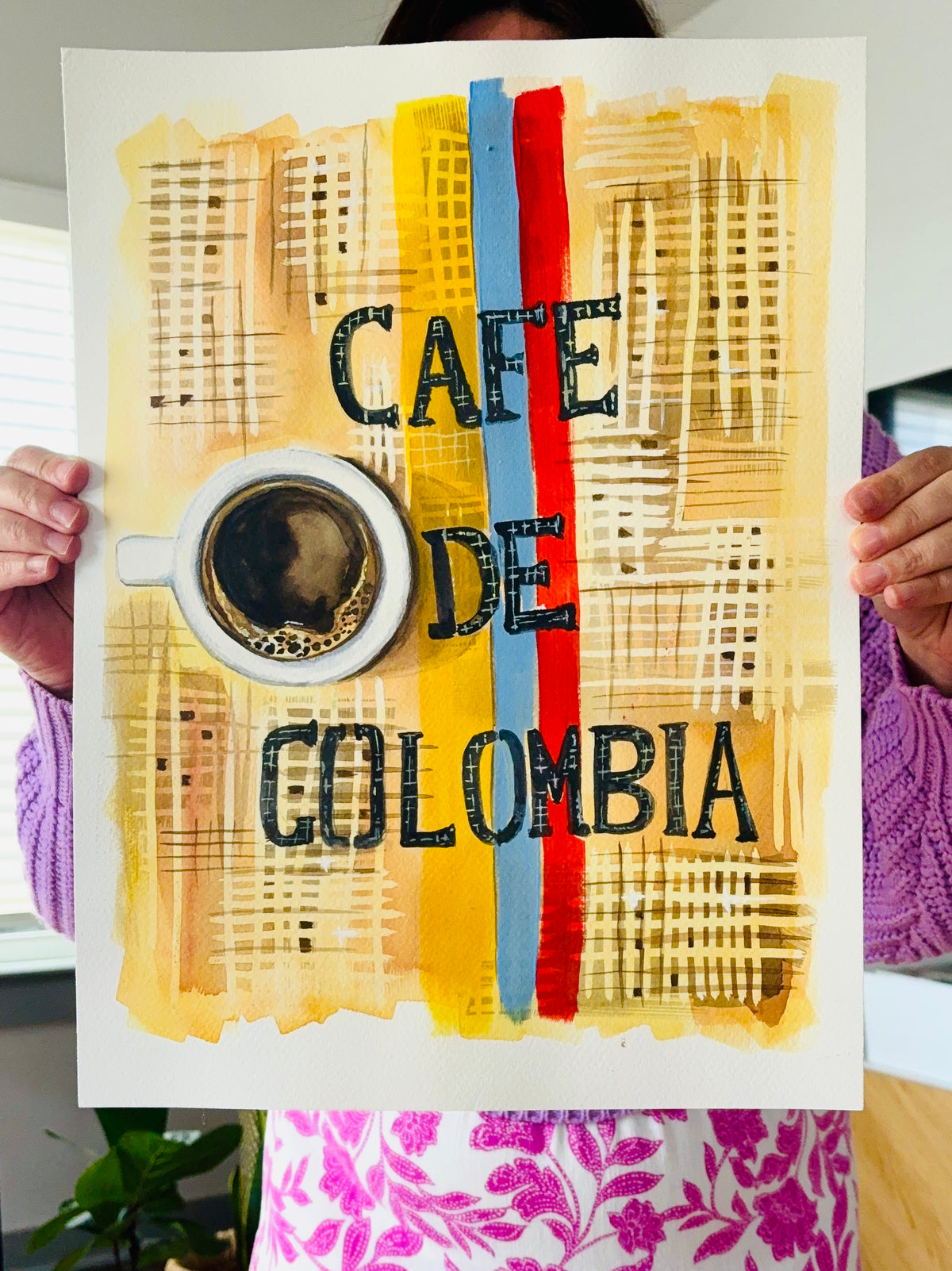 A person holds up a square art print featuring the words 'CAFE DE COLOMBIA' in black over a yellow, beige, and brown collage background with vertical blue and red stripes and a coffee cup illustration.