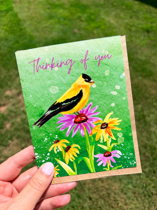 A hand holding a greeting card outdoors. The card features a watercolor-style yellow bird perched on purple and yellow flowers against a green background, with the handwritten text 'Thinking of you' at the top.