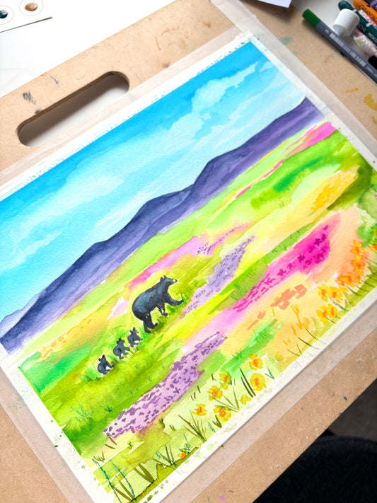 A colorful watercolor painting of a bear family in a grassy field with purple flowers and blue mountains, displayed on a wooden artist palette with paint markers nearby.