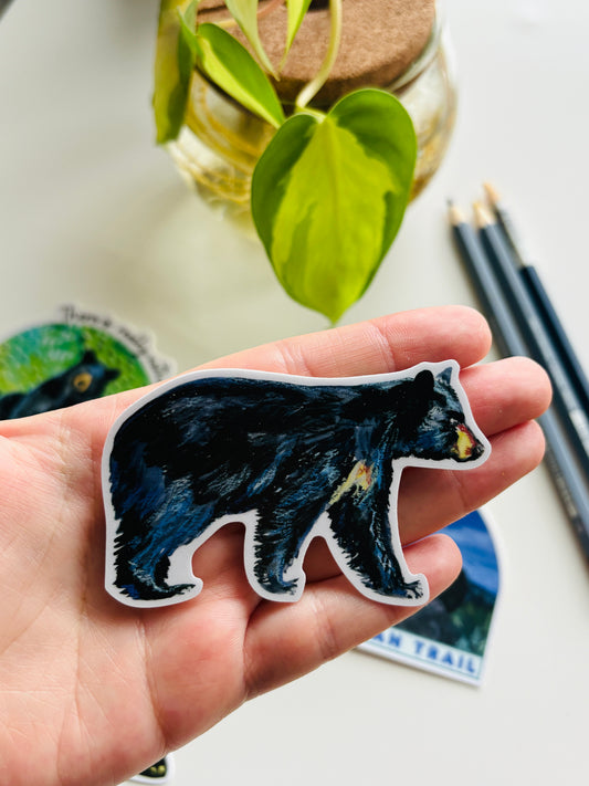 Hand holding a black bear sticker with a blurred background of plants and pencils