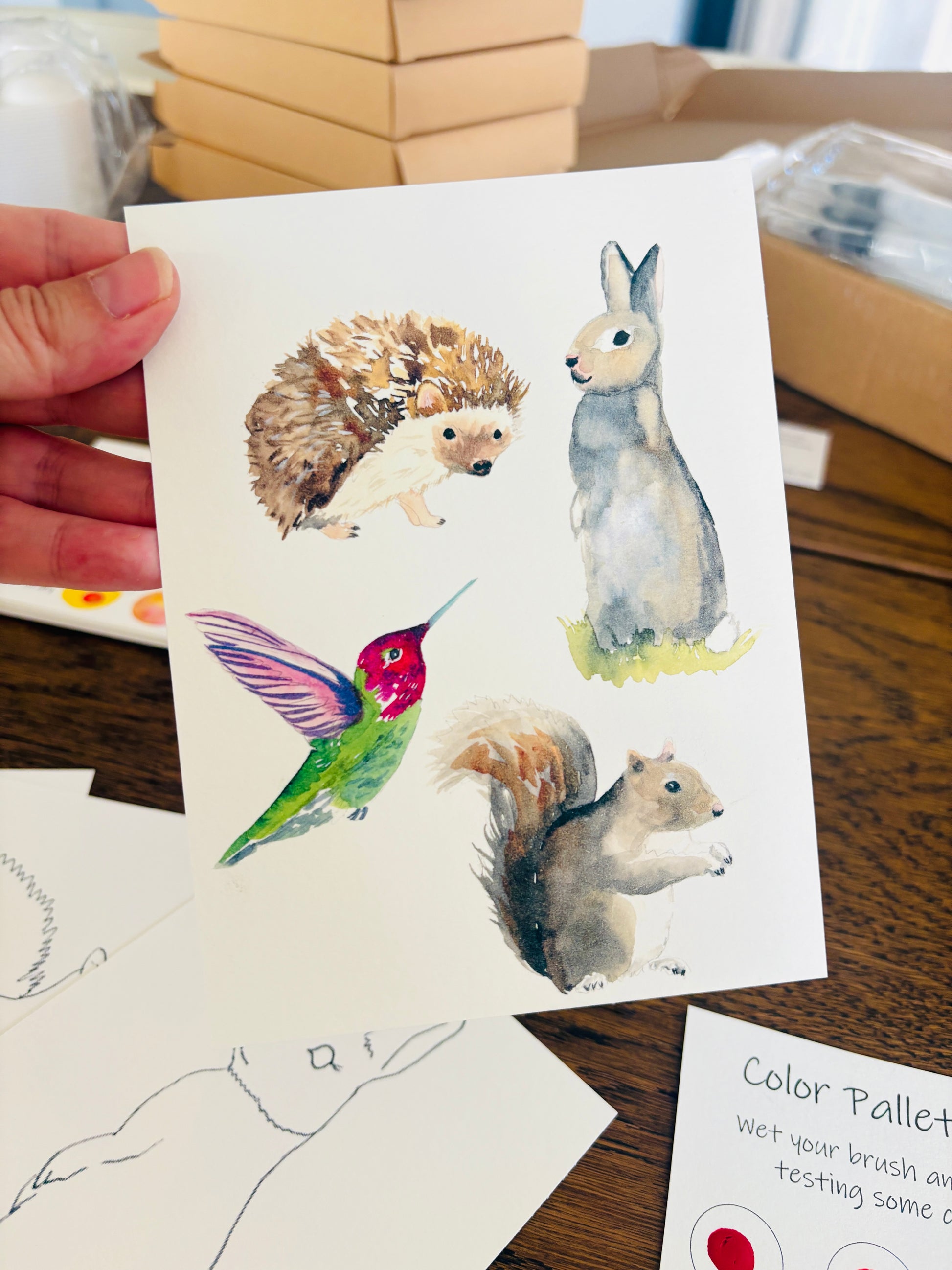 Hand holding a card with watercolor illustrations of animals on a wooden surface.