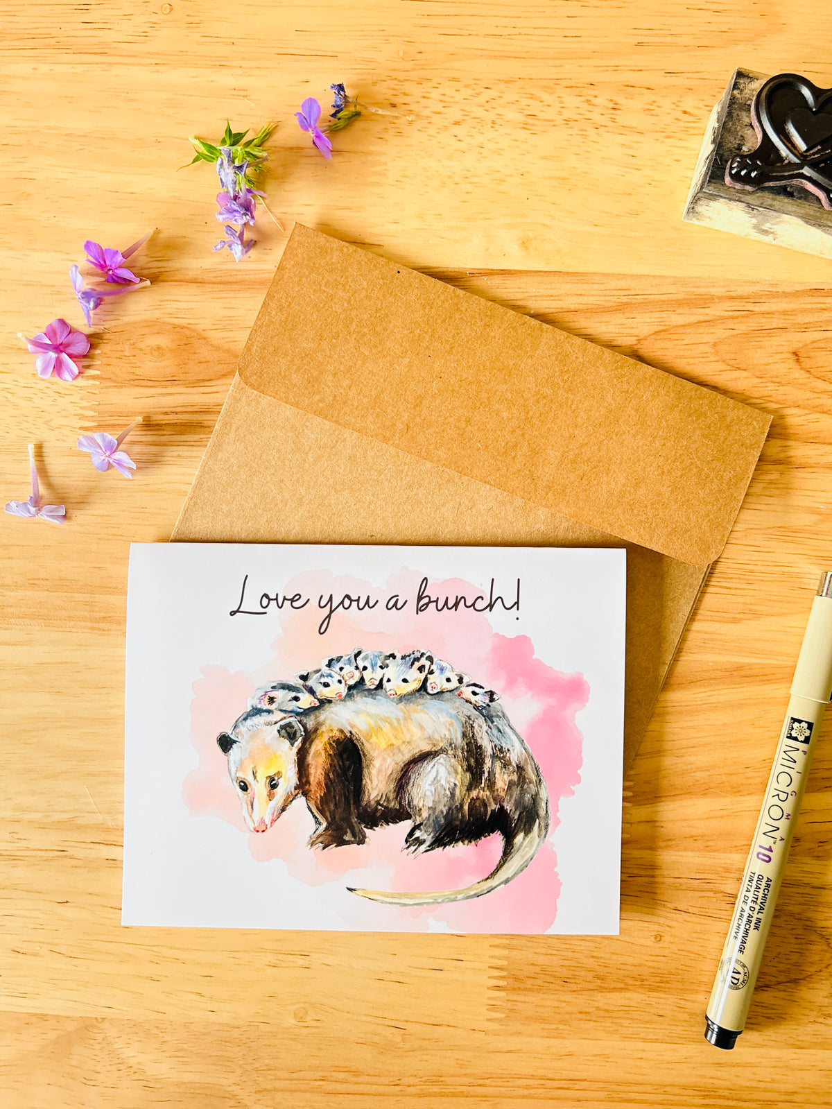 A greeting card with a watercolor illustration of an opossum on a pink watercolor splash, with the text 'Love you a bunch!' at the top. The card is displayed on a wooden table next to a brown kraft envelope, a pen, and small purple flowers.