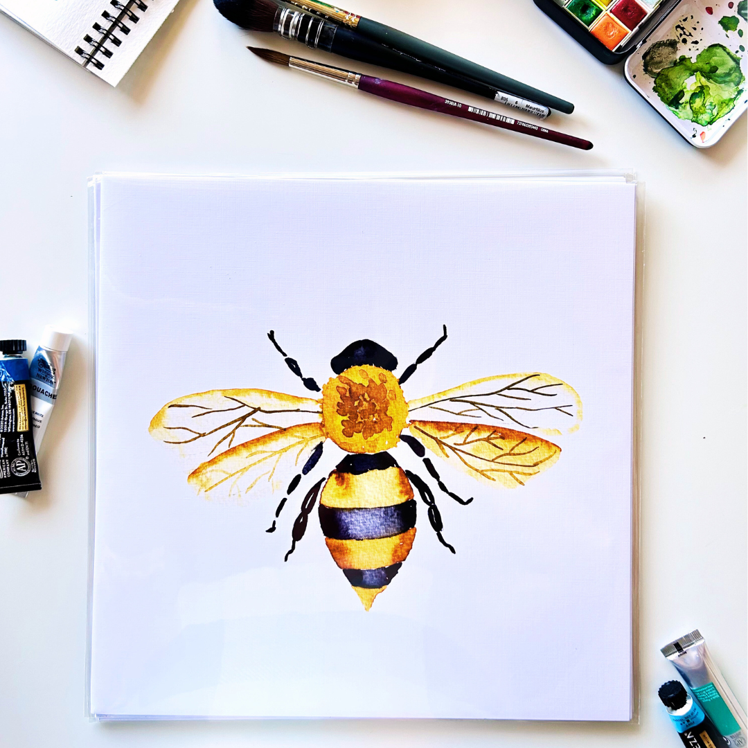 A framed art print of a watercolor painting featuring a detailed bee design, displayed on a table with art supplies and a sketchbook.