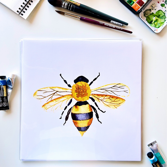 A framed art print of a watercolor painting featuring a detailed bee design, displayed on a table with art supplies and a sketchbook.