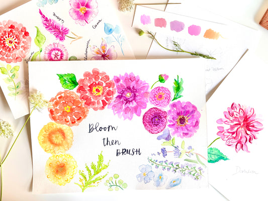 A styled flat-lay of watercolor floral art prints and swatches on white paper, featuring colorful painted flowers, green leaves, and the phrase 'Bloom then BRUSH' on the central print.