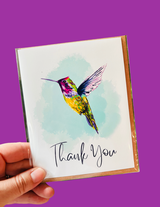 A hand holds a square greeting card with a watercolor-style hummingbird illustration and the words 'Thank You' in black script. The card has a white front, a gold-toned border, and is photographed against a solid purple background.