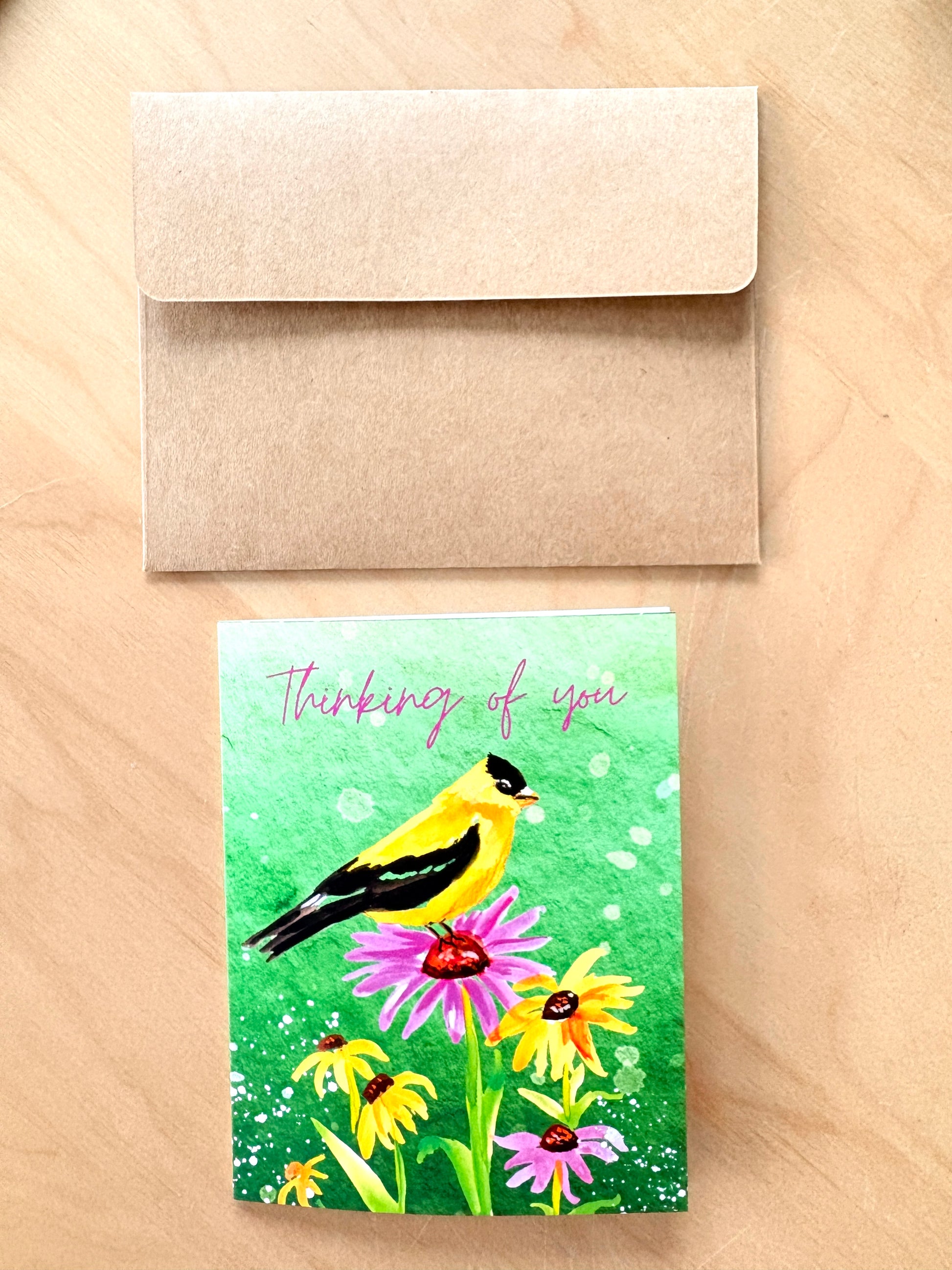 A square greeting card with a colorful illustration of a yellow and black bird perched on pink and yellow flowers against a green watercolor background, with the words 'Thinking of you' in pink script. The card is shown below a plain kraft paper envelope on a light wood surface.